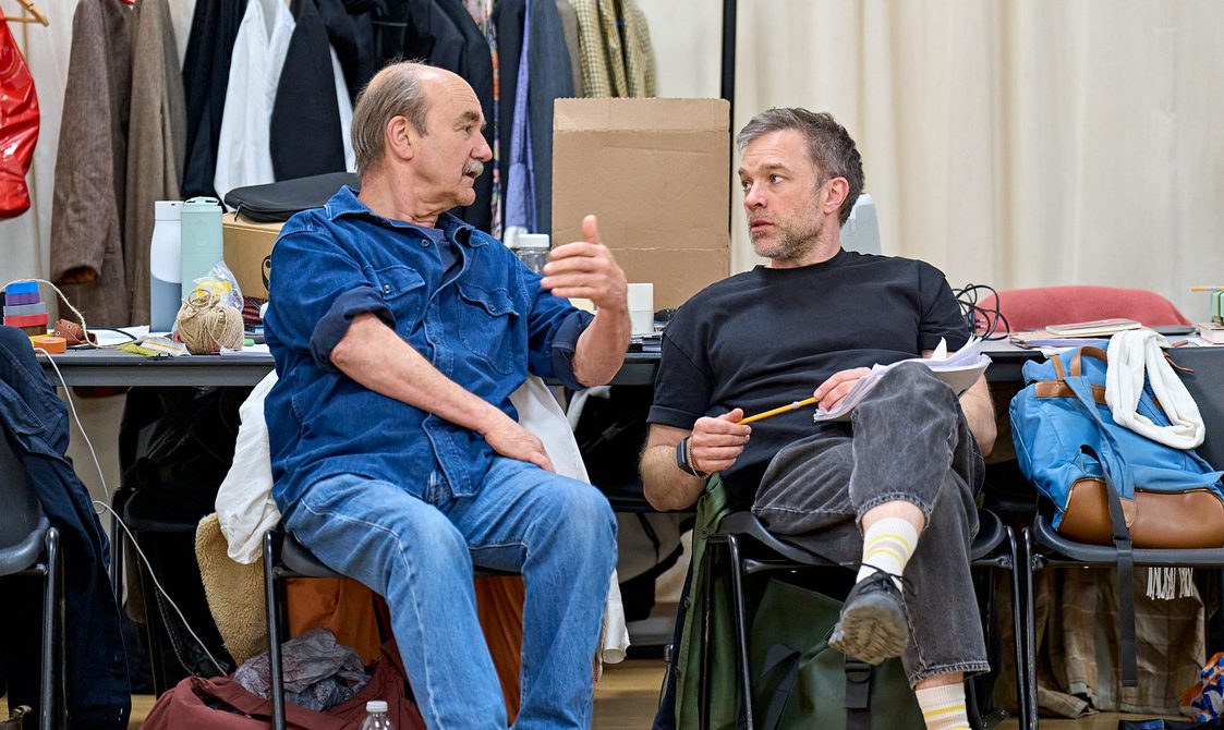Two men chat to each other sitting in a rehearsal room.