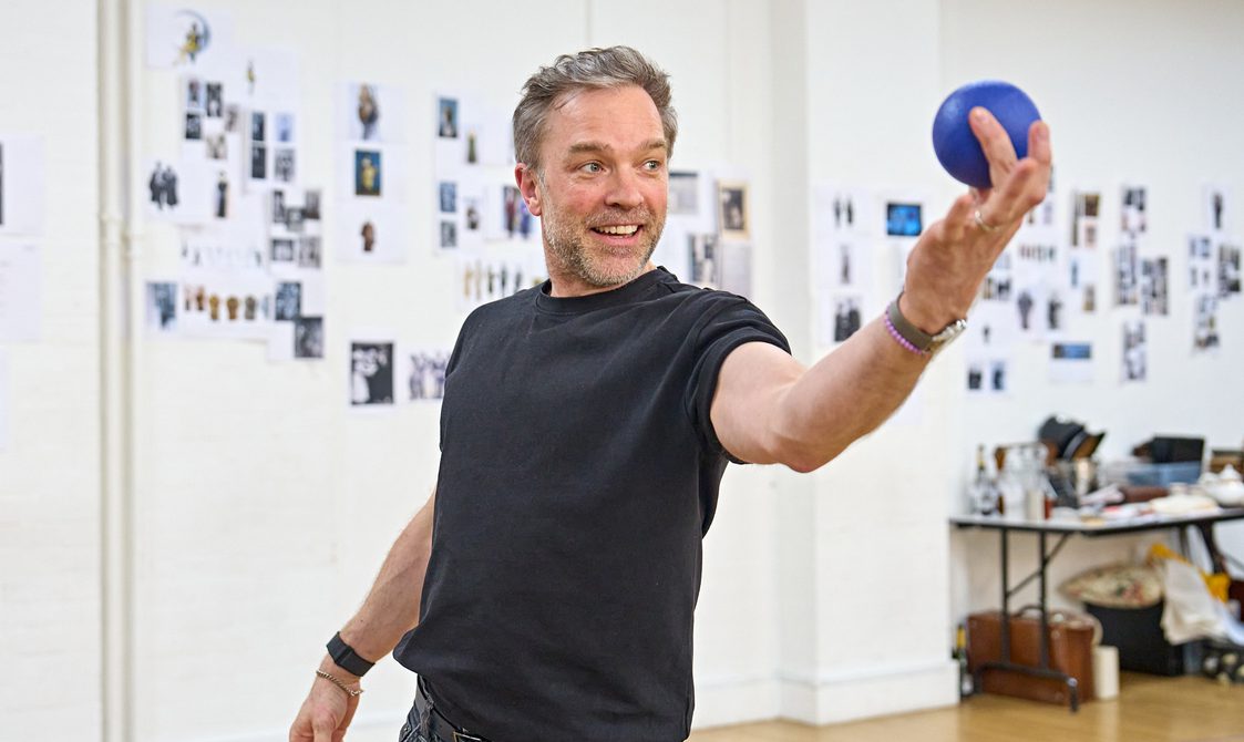 A man with light grey hair stands holding a blue ball in front of him.