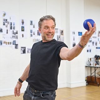 A man with light grey hair stands holding a blue ball in front of him.