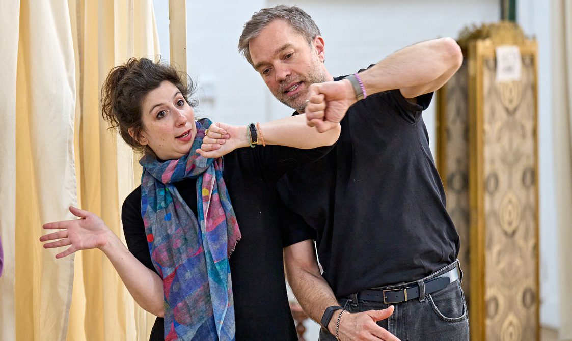 A woman with dark hair and a colourful scarf is leaning to the left with her arms swaying. Next to her is a man with light grey hair holding his arm out in front of him.
