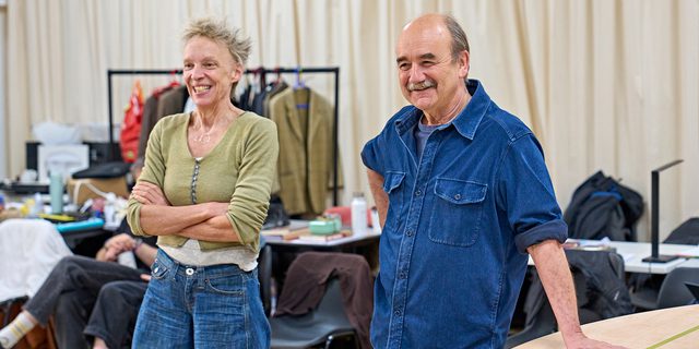 A woman with spiky blonde hair is stood with her arms crossed. Next to her is a man with a grey moustache leaning against a table. They are both smiling.