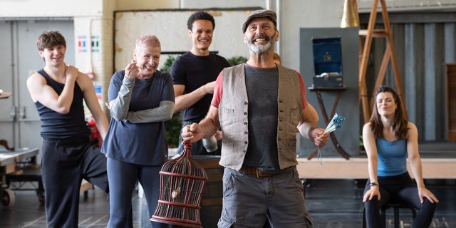 A performer with grey facial hair stands singing joyfully in a studio, holding a birdcage in one hand and a closed fan in the other. He is wearing grey cargo shorts, a grey and red t-shirt, a brown waistcoat and a flat cap. Behind him, a group of other performers sit and stand, watching on, leaning on a wooden barrel.