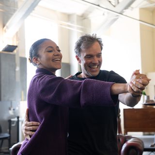 A younger woman with her dark, curly hair tied in a tight bun dances joyfully in hold with a man with grey hair in a studio. Both smile and look towards their hands which are clasped and held out straight ahead of them. They wear casual clothes.