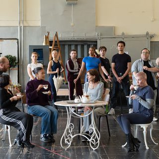 A group of performers in a rehearsal studio. In the centre, four women sit around an outdoor white café table holding traditional tea cups. The younger woman talks to the older woman in the middle, whilst the others sip their tea. Around them, a group watches on with interest, standing proudly.