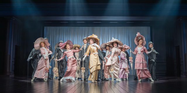 A company of performers stand together, dressed for Royal Ascot. The women have big fascinators on their heads, long satin dresses, silk gloves and parasols. The woman in the middle is wearing yellow and holds her hand under her chin. The men are behind them, all in coat tails. They are all posing. Four spotlights shine down on the group.