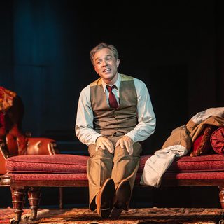 A man in a waistcoat and red tie sits on a red chaise lounge. His jacket has been thrown next to him. He stretches out his legs in front of him. A red arm chair is behind him and rugs cover the floor.