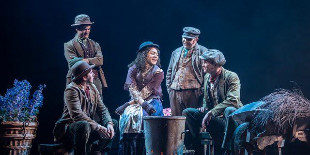 A woman with curly brown hair and a hat sits in the centre of four men. There is a metal bin, glowing orange from inside in front of them. A basket of flowers and a wheel barrow of straw are either side of the gorup. The men are looking and smiling at the women as she speaks to them. Two are sitting, the other two are standing and have their arms crossed and the other with his hands in his pockets.
