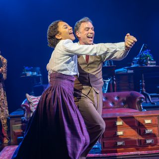 A man and woman dance together, wide smiles on their faces. A man in the background watches them, clapping his hands together. He is wearing a gold patterned house coat.
