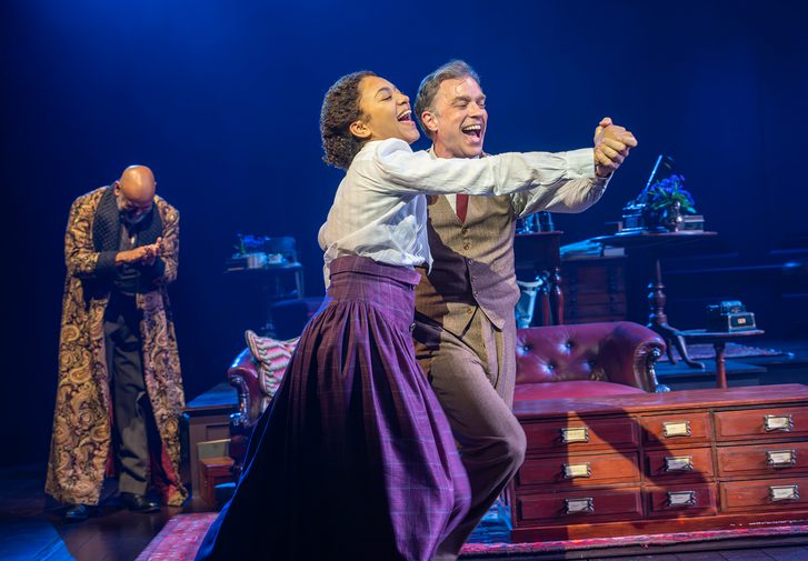A man and woman dance together, wide smiles on their faces. A man in the background watches them, clapping his hands together. He is wearing a gold patterned house coat.