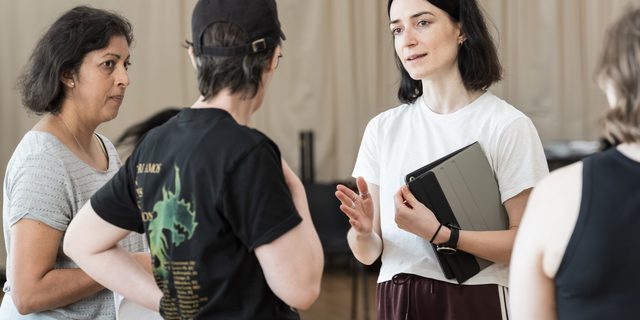 Four members of the company stand. The woman in the centre has short dark hair and is wearing a white t-shirt and burgundy tracksuit trousers. She is holding an iPad and talking to the performer in front of her, holding her hand out in front of her, directing. The performers on the left are looking at her. One is wearing a black cap and black graphic t-shirt. The other is wearing a grey t-shirt and jeans. On the right is another member of the company with short brown hair and wearing a black tank top.