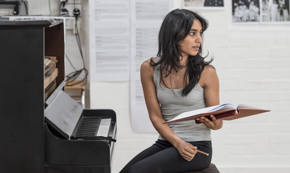 A woman with long dark hair, a grey vest-top and black leggings sits in front of a piano. Her body is turned away from the piano and holds an open book in her left and and a pen in her right.