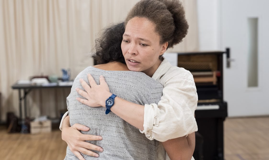 Two women embrace in a hug. The woman facing the camera has her hair in a bun and is wearing a white shirt with the sleeves rolled up and a blue wristwatch. The other woman has her head on her shoulder. Behind them is a rehearsal studio and a piano.