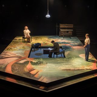Three actors stand and sit around a pentagon shaped stage. There is a sunken square in the middle where an actor sits on a bench. They have very short brown hair and wear a grey suit. The actor is looking down at some papers. The woman on the left is also sat on a bench. She has short blonde wavy hair and wears a white shirt, beige trousers and has a scarf wrapped around her shoulders. She is sat up straight. The woman on the right wears a white blouse, blue trousers and her brown hair in a bun. She has her hands together in front of her as she looks down. A piano sits in the corner of the stage and a single lamp hangs from the ceiling. The floor is washed in colour and pattern.
