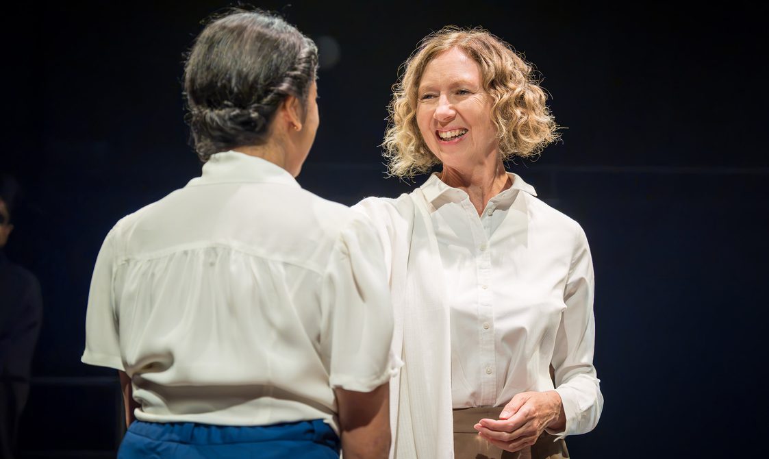 Two women face each other. The woman with her back to the camera is wearing a white blouse, blue trousers and her dark hair is twisted into a low bun. The other woman has short blonde hair that has been curled. She is wearing a white shirt and beige trousers. She is smiling widely, her right arm reaching out towards the other woman.