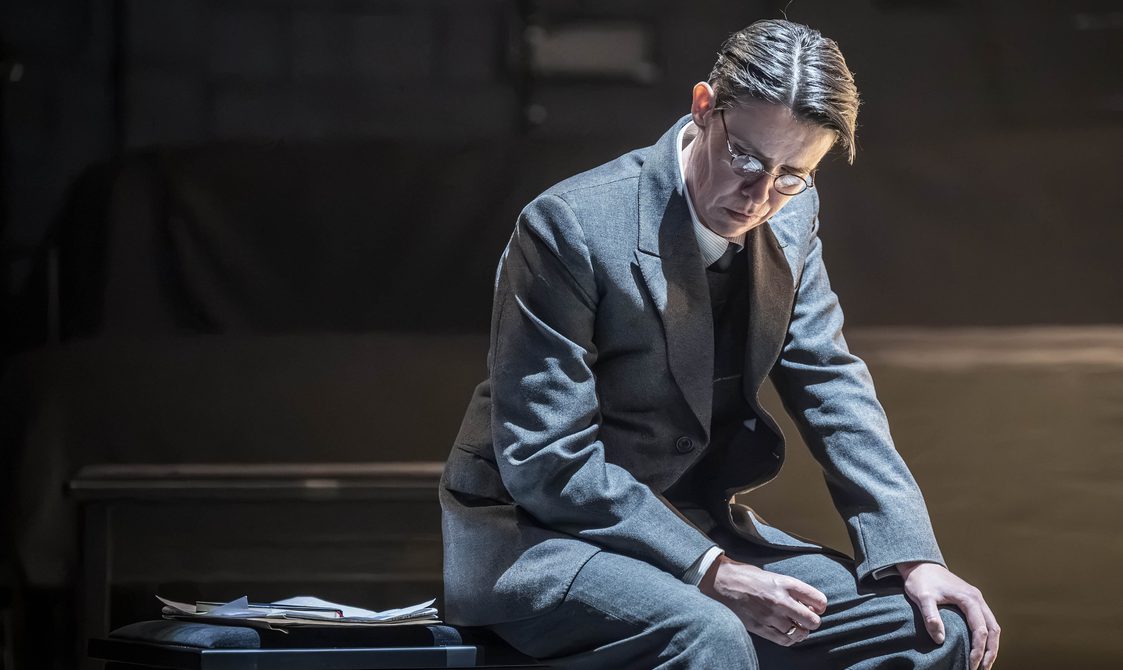 An actor sits on a bench. She has very short cropped brown hair parted to the side and round glasses. She is wearing a grey suit and a gold ring. She is looking down at the ground in defeat. A clipboard sits on the bench beside her.