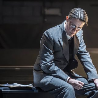 An actor sits on a bench. She has very short cropped brown hair parted to the side and round glasses. She is wearing a grey suit and a gold ring. She is looking down at the ground in defeat. A clipboard sits on the bench beside her.