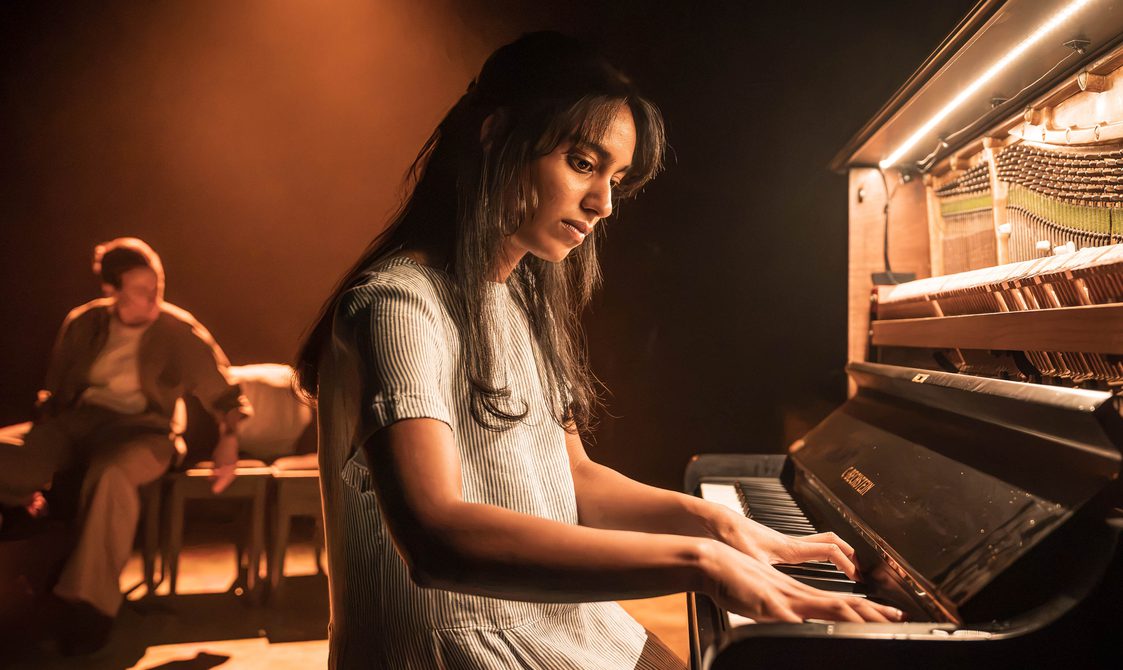 A woman with long dark hair and a pinstriped short sleeve dress sits at a piano. She has her fingers on the keys and looks down at them. Behind is an actor, blurred from view. They have their sleeves rolled up and are sat down. The stage is warmly lit and the piano is lit up from within.