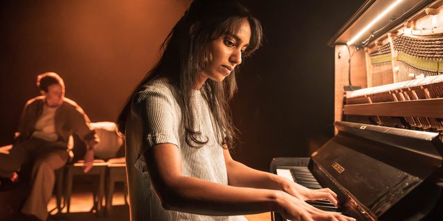 A woman with long dark hair and a pinstriped short sleeve dress sits at a piano. She has her fingers on the keys and looks down at them. Behind is an actor, blurred from view. They have their sleeves rolled up and are sat down. The stage is warmly lit and the piano is lit up from within.