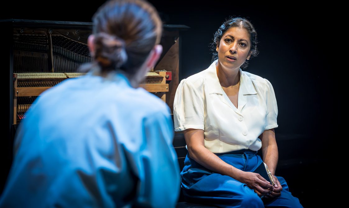 Two women are sat facing each other. The woman facing the camera has a white blouse, blue trousers and her dark brown hair twisted into a low bun. She has her hands in her lap, holding a small notebook. The woman with her back to the camera has her brown hair tied into a sleek low bun and wears a blue shirt. Behind them is a piano.