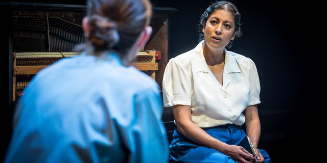 Two women are sat facing each other. The woman facing the camera has a white blouse, blue trousers and her dark brown hair twisted into a low bun. She has her hands in her lap, holding a small notebook. The woman with her back to the camera has her brown hair tied into a sleek low bun and wears a blue shirt. Behind them is a piano.