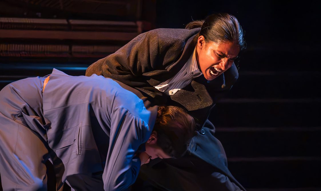 Two women are in a physical altercation. The woman on the right has her dark hair pulled into a low ponytail and has a long dark grey trench coat and striped shirt on. She is bent over, dragging the second woman, who is wearing a bright blue suit, down towards the floor. Her mouth is wide open and nose and eyebrows scrunched up as if she were yelling.