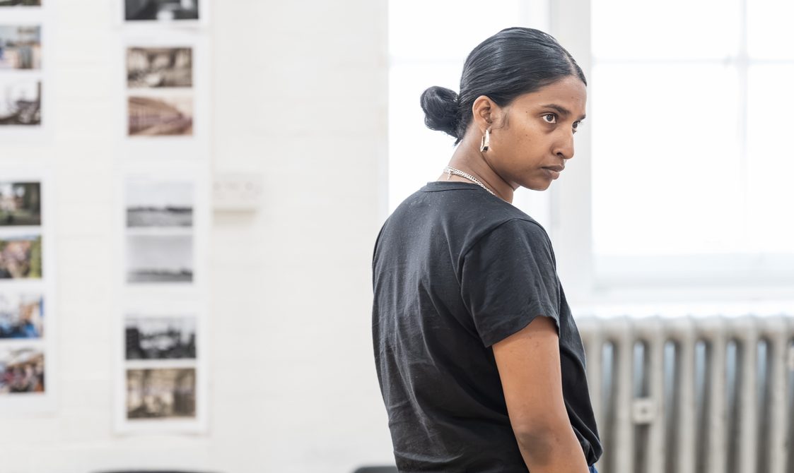 A woman with dark hair in a low bun and chunky hoop earrings has her back to the camera. Her head is turned to the side and has a serious expression on her face.