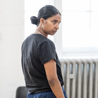 A woman with dark hair in a low bun and chunky hoop earrings has her back to the camera. Her head is turned to the side and has a serious expression on her face.