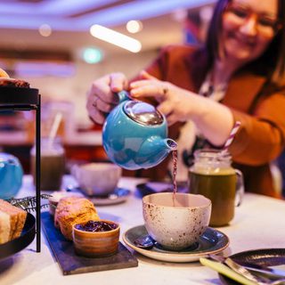 A woman pours tea into a cup; the table is full of delicious looking sandwiches, scones and treats.