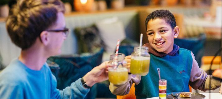 Two boys clink their glasses together at a table; they are surrounded by sweet treats on the table.