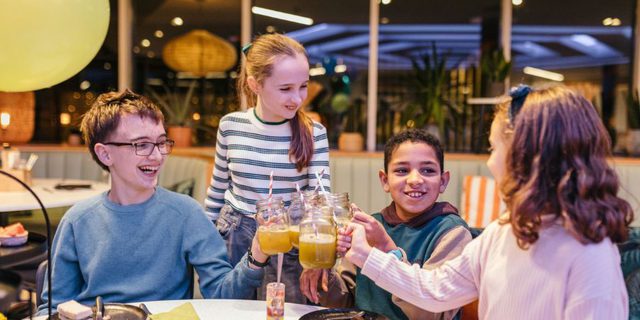 Four children clink their glasses together at a table; they are surrounded by sweet treats and balloons on the table.