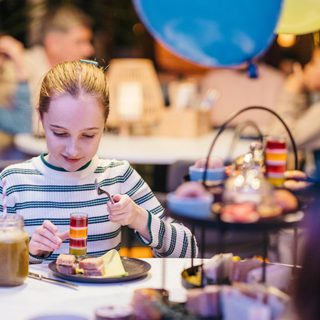 A girl in a stripy top eats a stripy jelly; she is surrounded by sweet treats - balloons and people are in the background.