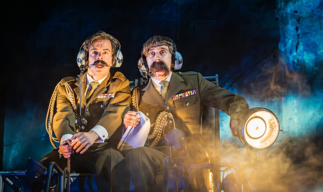 Two men sat next to each other wearing military uniform, both with dark short hair and moustaches and with shocked expressions on their faces. One of the men has a considerably larger moustache and is holding a map and large torch