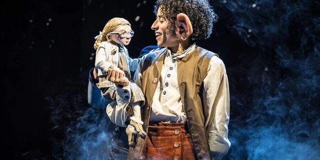 A man with short curly dark hair and big ears wearing a white shirt, orange/brown trousers and waistcoat, holding a small puppet of a girl