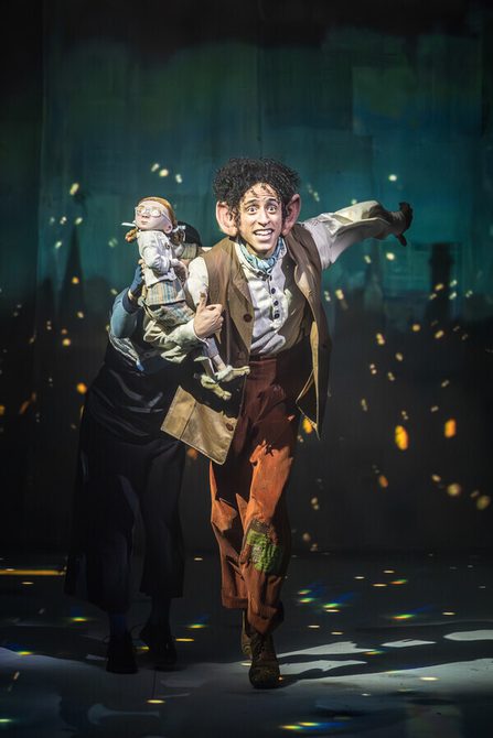 A man with dark brown curly hair and big ears wearing a white shirt and orange/brown trousers with a white shirt and brown waistcoat holding a small puppet on his arm in a running pose