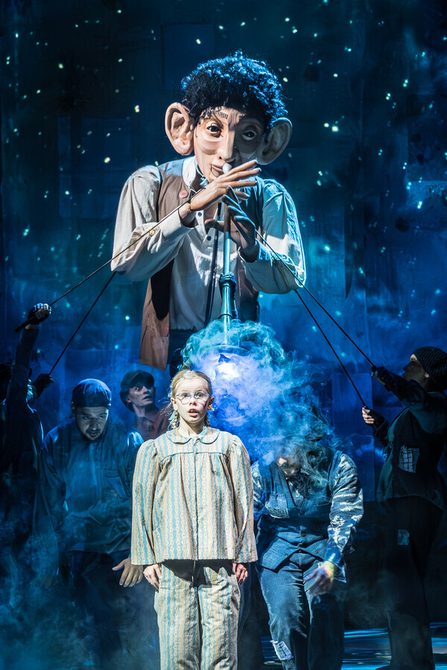 A giant puppet with short dark curly hair wearing a white shirt with a brown waistcoat on holding a big pipe to his lips. In front of the puppet there is a girl with stripped pyjamas on with a shocked expression on her face