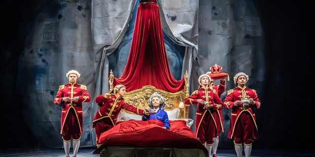 A woman with grey hair in purple pyjamas lying in a bed with a red duvet and gold-plated headrest holding a cup of tea. Next to her are five people in servant uniform