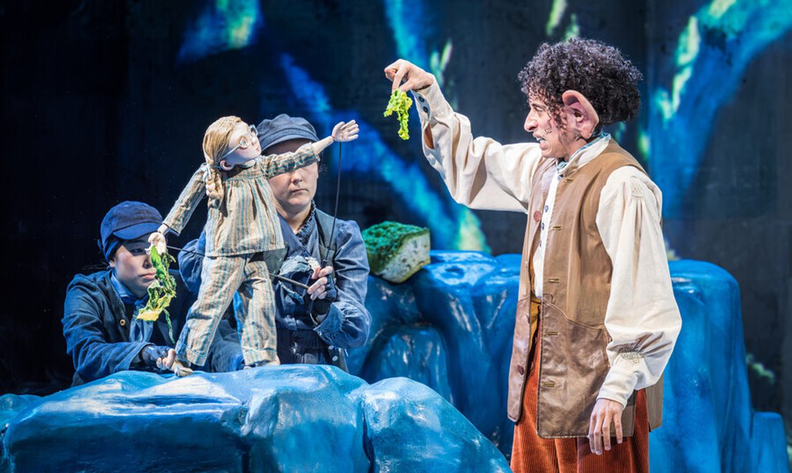 A man with dark curly hair and big ears wearing a white shirt, brown waistcoat and orange trousers holding up a green piece of fabric in front of a small girl puppet wearing striped pyjamas. The puppet is being operated by two women wearing dark clothes and a hat