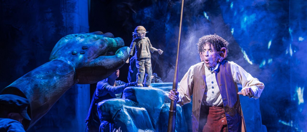 A big puppet with spiky hair on the ground holding up his big arm towards a man with short curly hair wearing a white shirt, waistcoat and orange/brown trousers looking quite shocked holding up a long pipe. In the background a small puppet is standing on a large rock