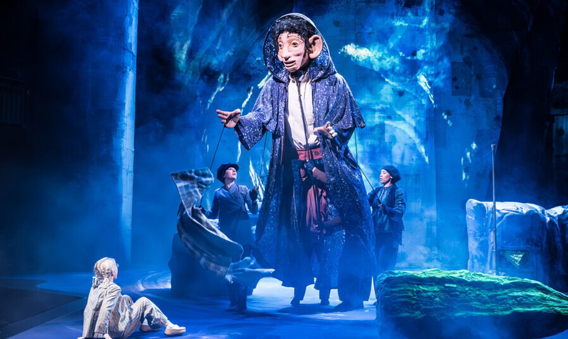 A small girl with blonde hair wearing stripped pyjamas is sat down on the ground looking at a giant man puppet that is wearing a long purple cloak being operated by two women in dark clothes and hats