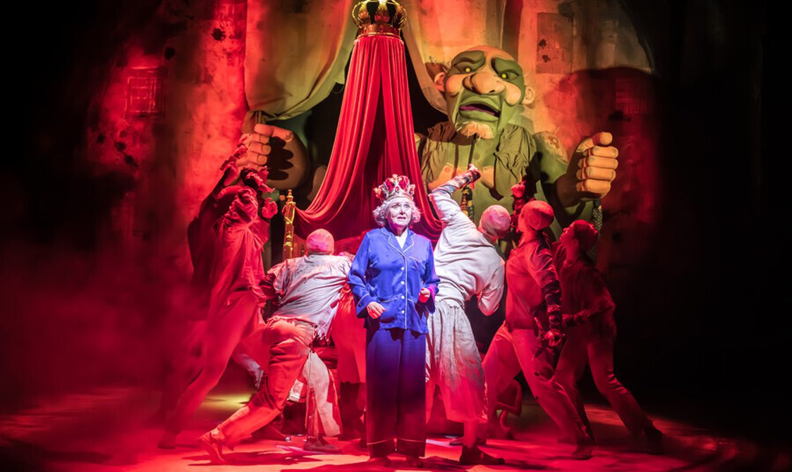 A woman wearing long purple pyjamas with grey shirt hair and a crown on her head stood in front of a large puppet and a group of people dressed in white clothes