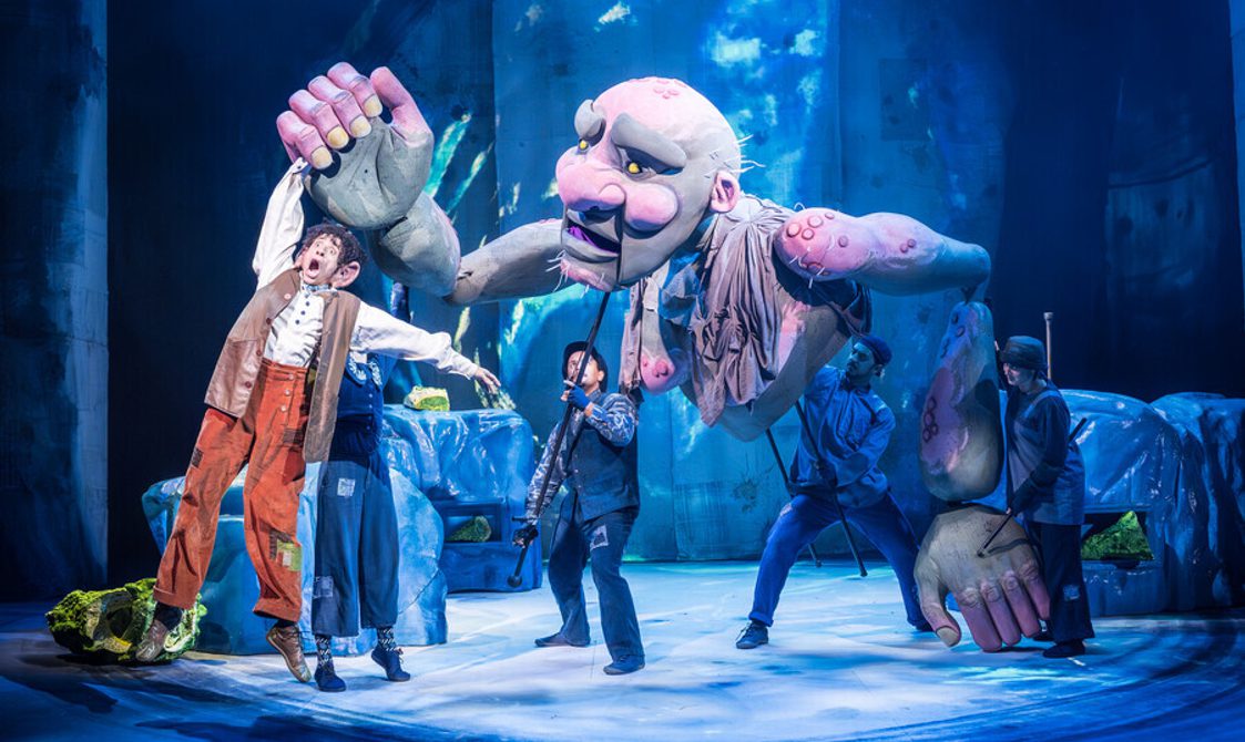 A man with curly black hair wearing orange/brown trousers and a white shirt and brown long waistcoat being held up in the air by a giant green and pink puppet. Four people in dark clothes and hats are stood below operating the puppet