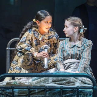A young girl with dark brown long hair wearing yellow and blue pyjamas holding up a torch to her face. She is sat talking on a bed next to a girl with light brown hair who is wearing green and brown stripped pyjamas