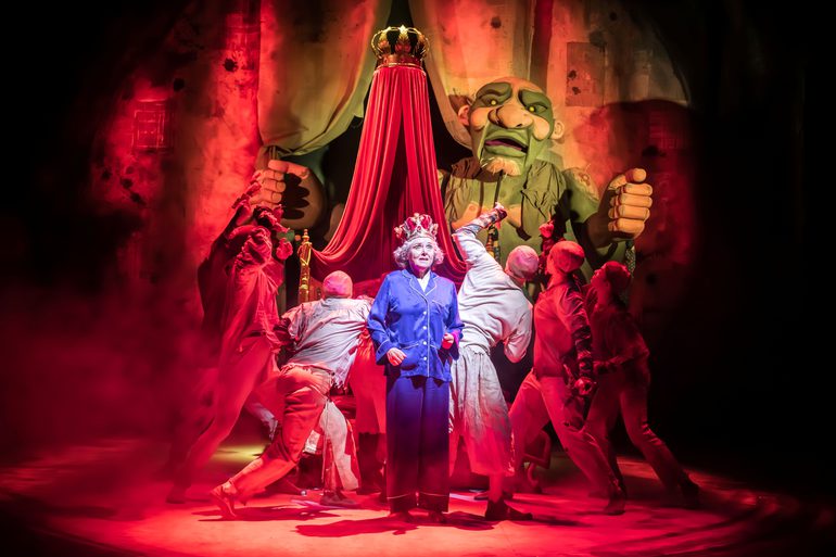 A woman wearing long purple pyjamas with grey shirt hair and a crown on her head stood in front of a large puppet and a group of people dressed in white clothes