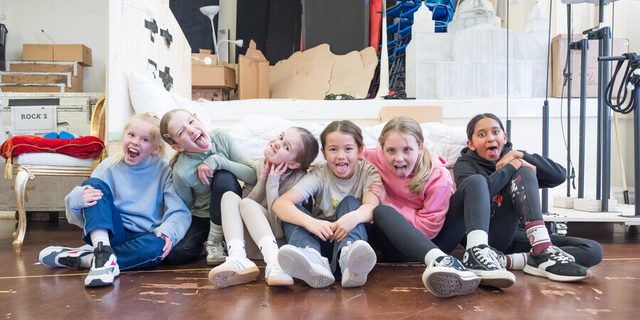 Six young girls sit in a line on the floor, all mischievously gurning for the camera with their tongues out or pulling funny faces.