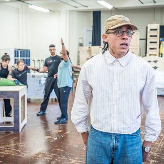 A man wearing a white striped shirt, jeans, beige baseball cap and glasses is standing looking out with a startled expression. Behind him, out of of focus, four people are standing in various attitudes, one with his arm raised.