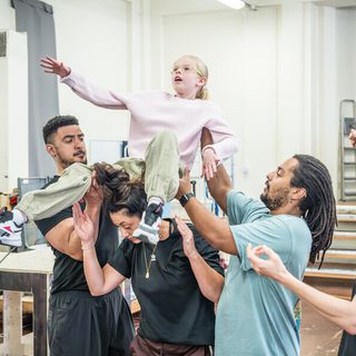 A young girl with blonde hair and glasses, wearing a pink top, is held aloft above the heads of four people. She is looking up with a look of amazement.