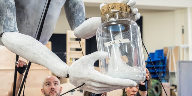 The arms and lower torse of a giant puppet is being held up and manipulated by some puppeteers. The puppet is holding a large glass jar with a cork lid; it has a label reading 'Jar A' and some white gauze is inside.