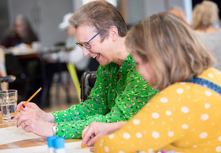 Two older women sit at a desk writing and laughing together.
