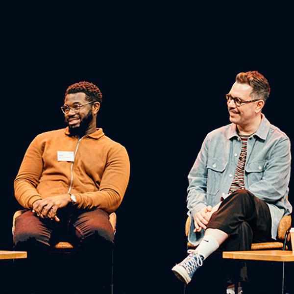 Three men and one woman, of different ages, dressed in casual clothes are sat on our stage holding microphones. One person is talking into the microphone while the others listen