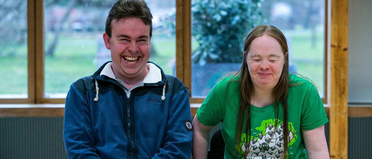 A man and woman are sat down in the Pimlott building, smiling and laughing. He is on the left and wearing a blue coat. She is on the right and is wearing a green t-shirt with sheep on it. Behind them you can see Oaklands Park.